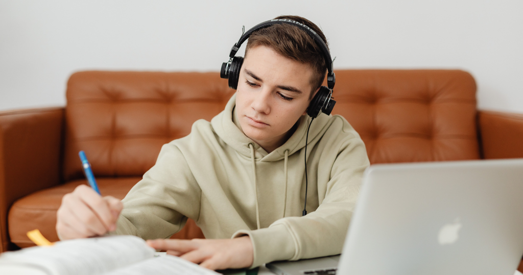 Joven estudiante con audífonos escribiendo en un cuaderno frente a una laptop